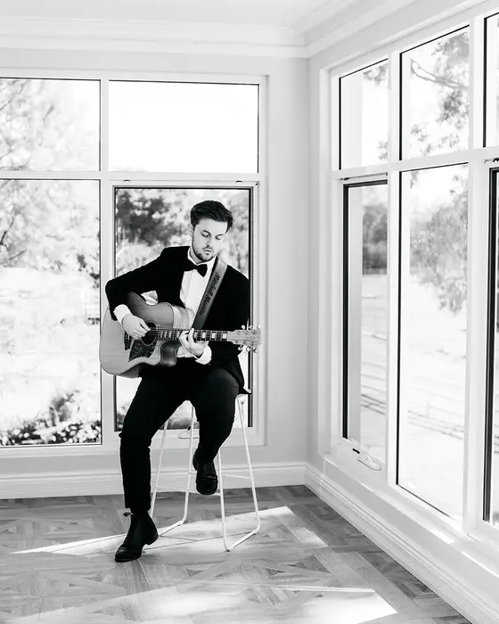 Mitchell playing acoustic guitar beside tall windows